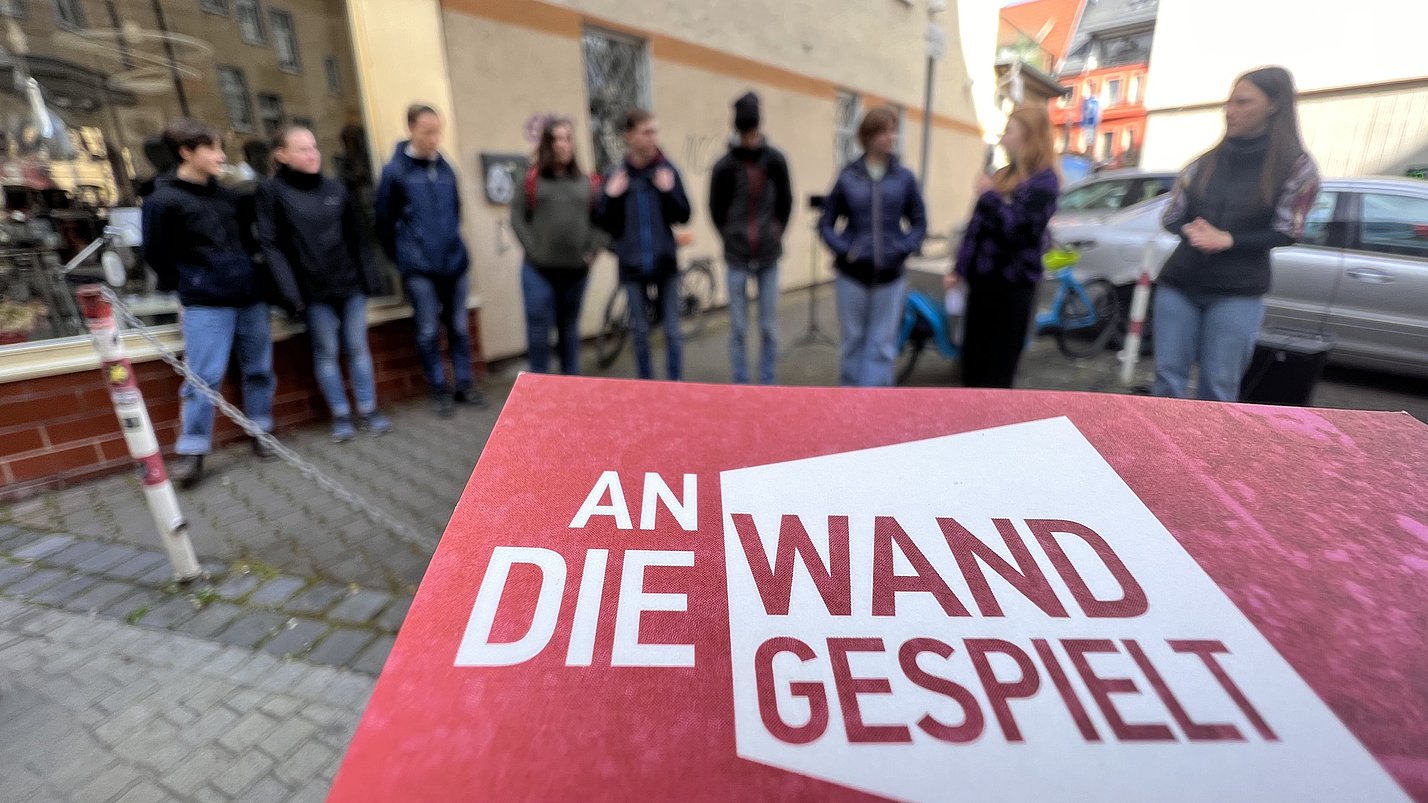 Flyer des Projekts "An die Wand gespielt". Im Hintergrund stehen Menschen in einem Halbkreis.