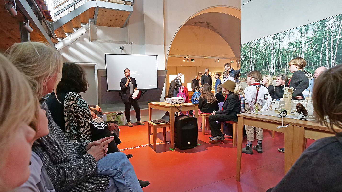 EIn Mann spricht in ein Mikrofon und viele Kinder hören ihm zu.