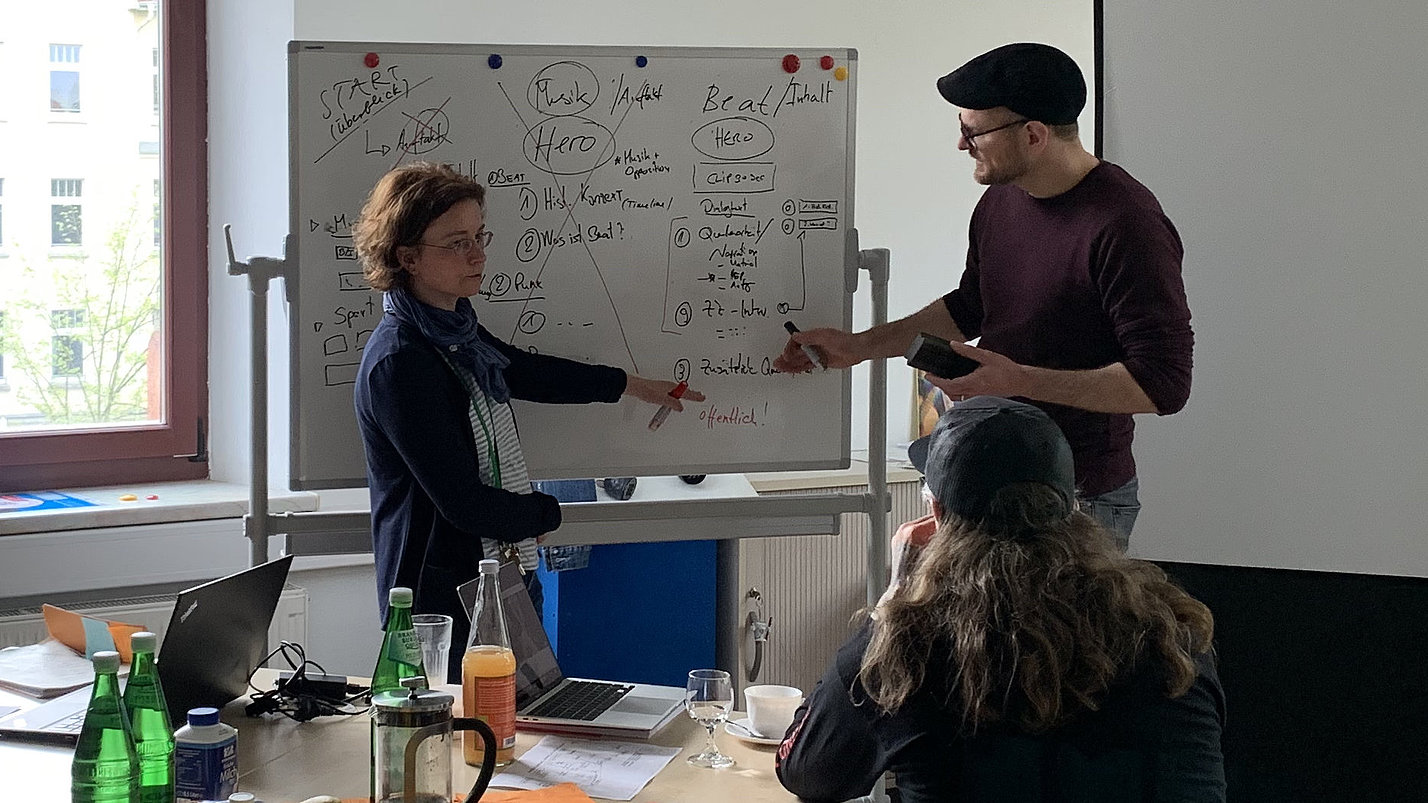 Zwei Menschen malen etwas an eine Tafel. Eine weitere Person sieht ihnen zu.