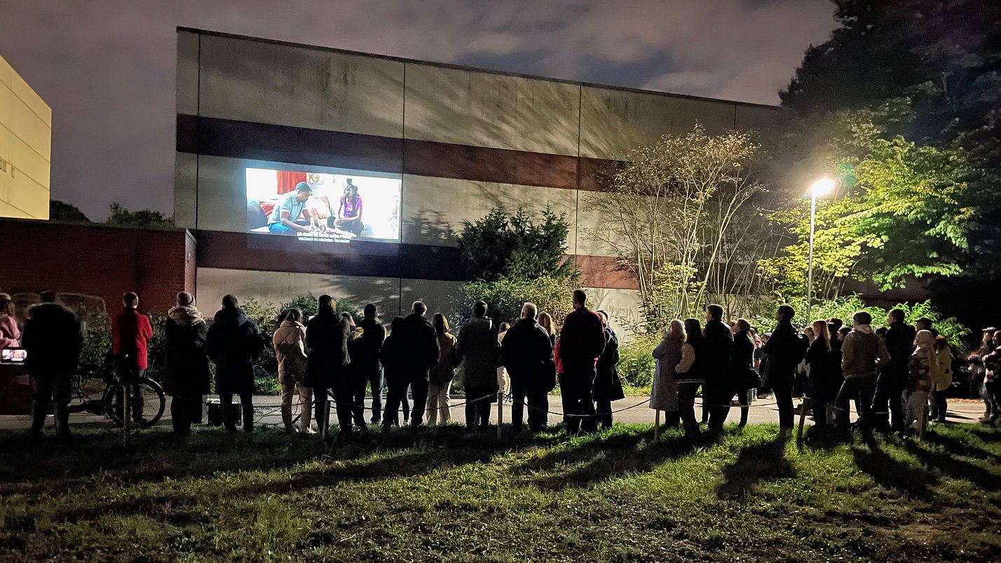 Viele Menschen stehen um ein Gebäude an dessen Fassade ein Video projiziert wird.