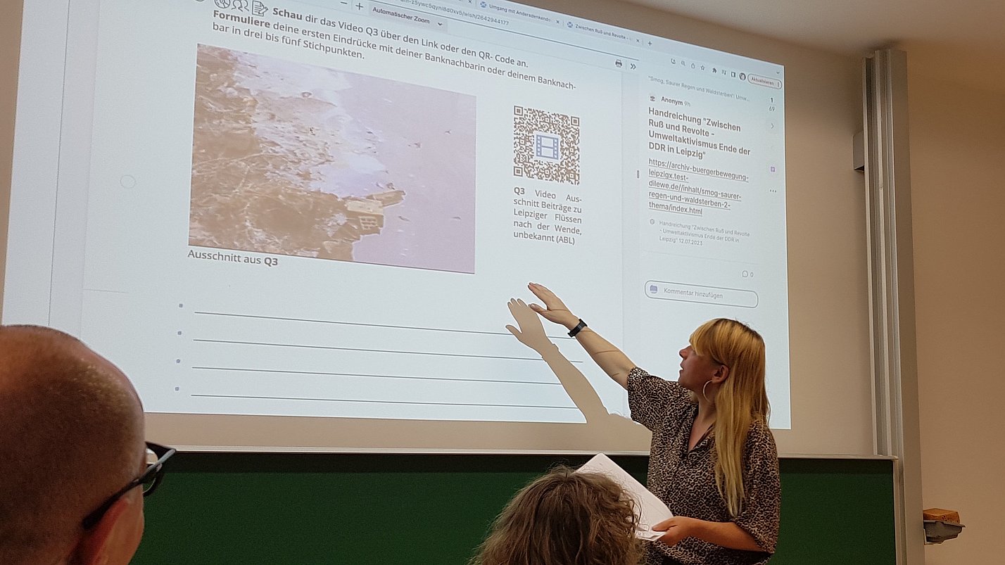 Eine Frau erklärt etwas an einer Präsentation. Weitere Menschen hören ihr zu.