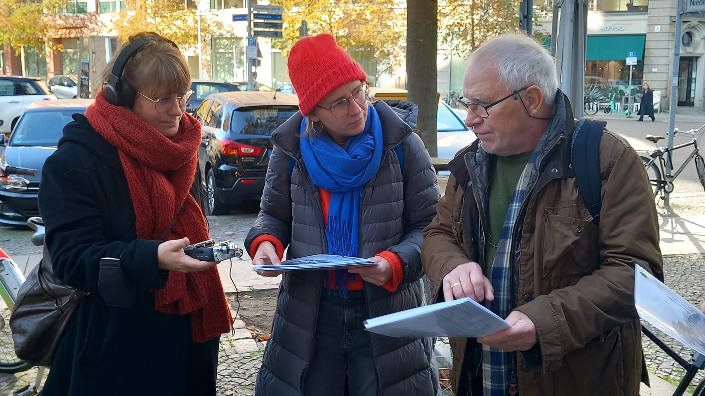 Drei Personen stehen mit Zetteln in der Hand draußen und unterhalten sich.