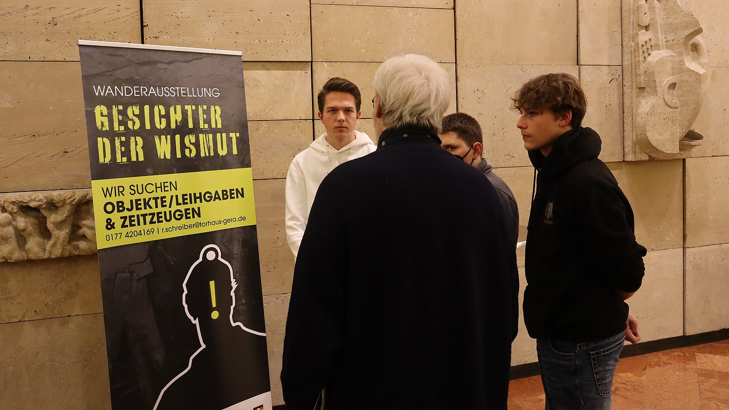 Drei Jugendliche stehen neben einem Banner des Projekts und sprechen mit einem älteren Mann.