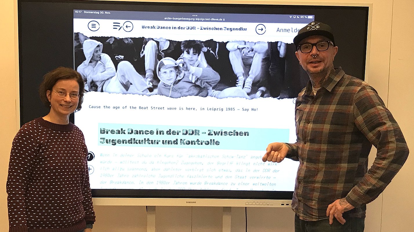 Zwei Personen stehen vor einem Bildschirm auf dem eine Präsentation zu "Breakdance in der DDR" gezeigt wird.