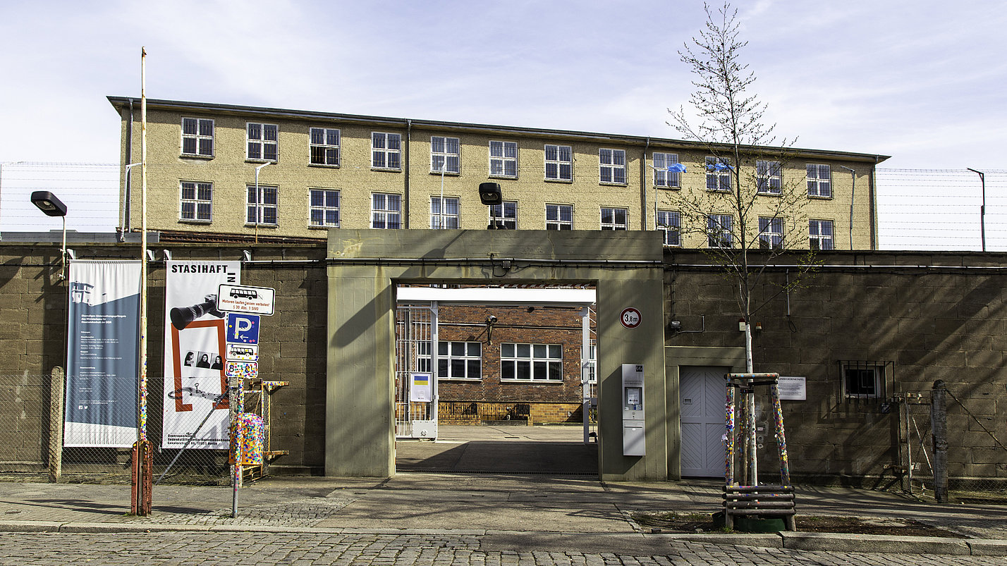 Ein in eine Gefängnismauer eingelassenes Eingangstor mit einem dahinter hervorragenden gelben Gebäude.