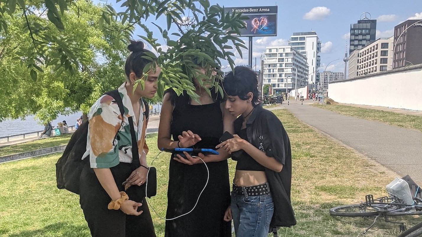 Drei Jugendliche stehen auf einer Wiese und schauen auf ein Tablet.